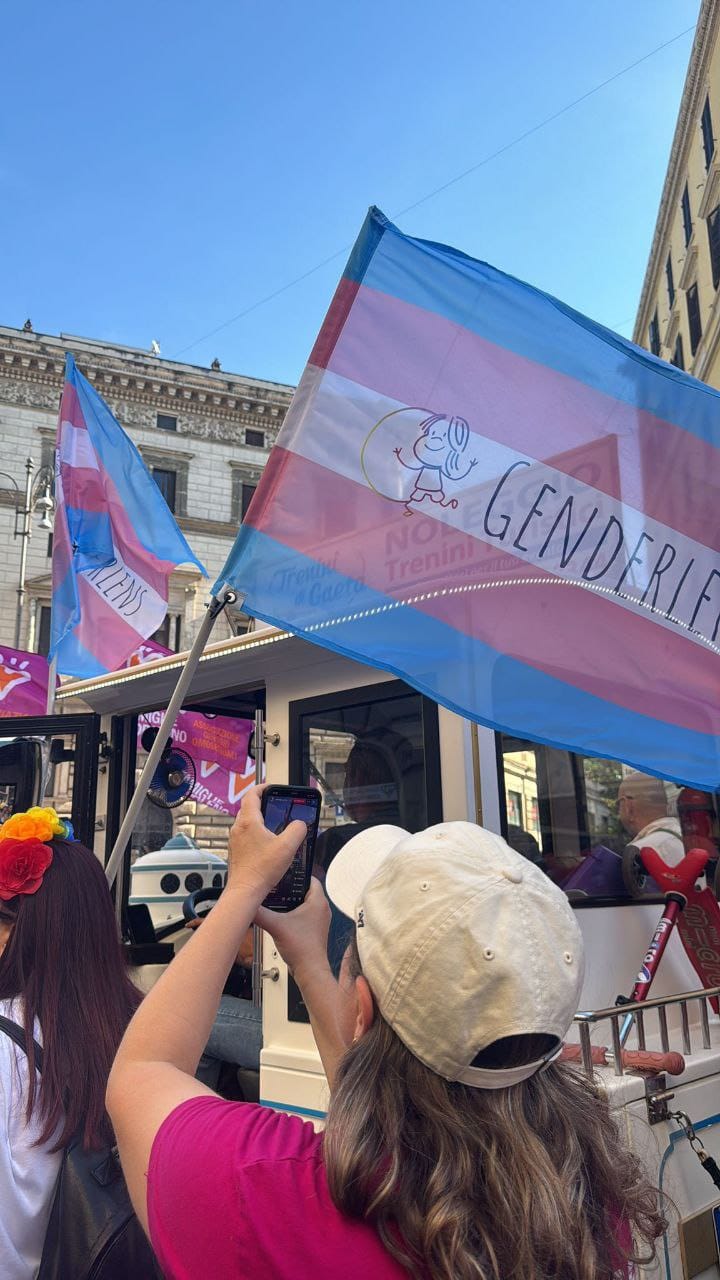 Striscione con bandiera trans e logo GenderLens sventolato da persone di spalle durante il pride.