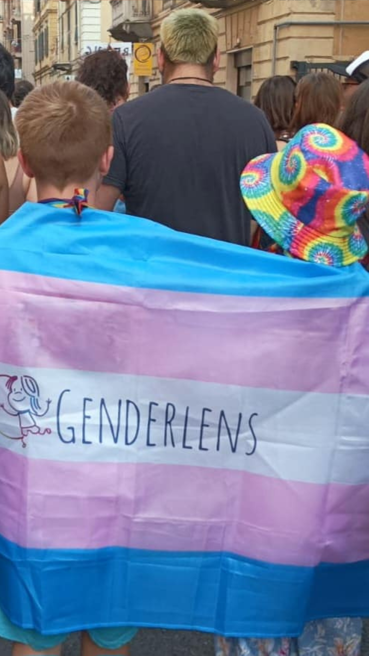 Striscione con bandiera trans e logo GenderLens tenuto come mantello da persone bambine con cappelli arcobaleno e outfit colorati, al pride.