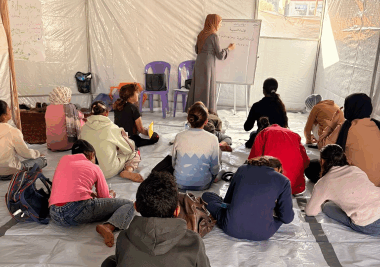 Lezione in tenda palestinese: persona in abito lungo di spalle scrive alla lavagna bianca mentre persone bambine sedute per terra di spalle ascoltano attente.